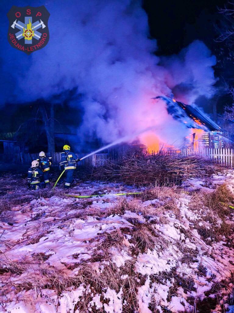 Strażacy w mundurach gaszą pożar w nocy, otoczeni dymem i płomieniami, w pobliżu drewnianego ogrodzenia.