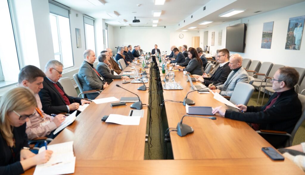 Grupa mężczyzn i kobiet w formalnych strojach uczestniczy w spotkaniu przy długim stole w biurze.