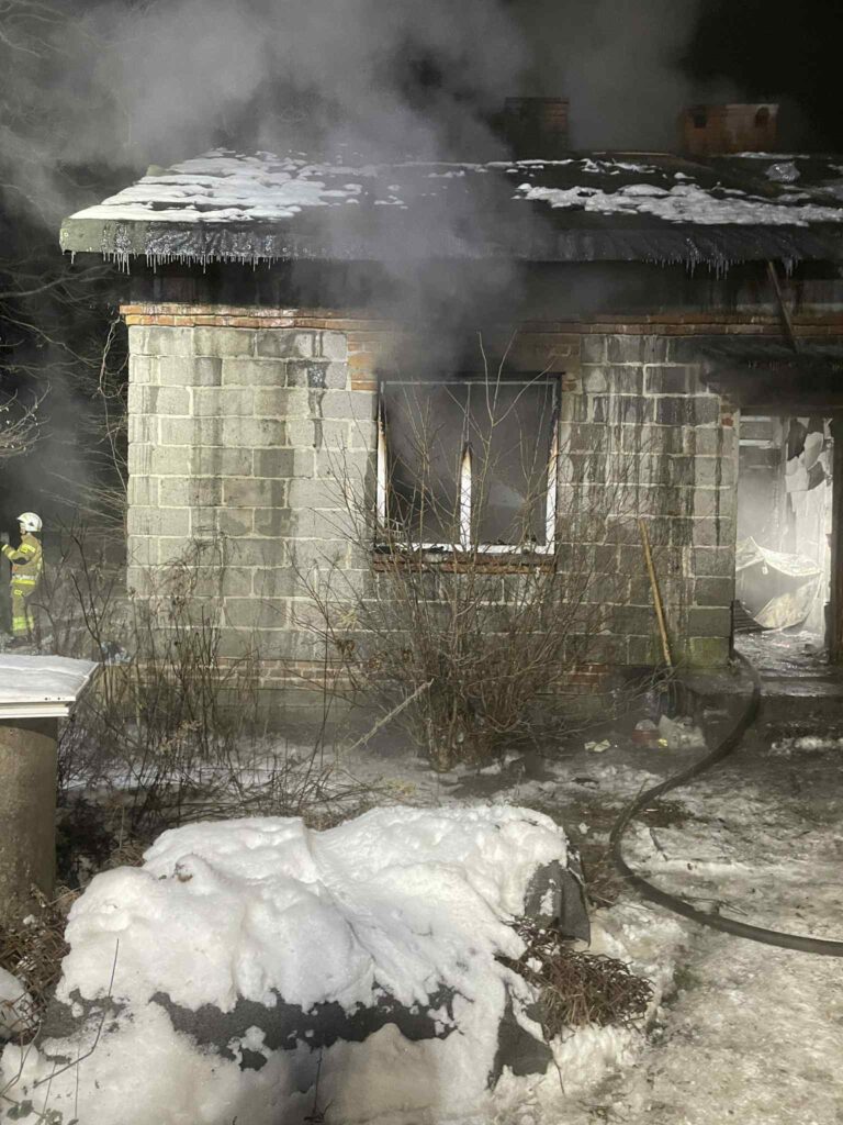 Z budynku wydobywa się dym, a w tle widoczny jest strażak w sprzęcie ochronnym, otoczony śniegiem.