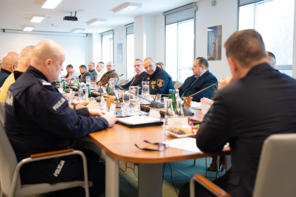 Grupa mężczyzn w garniturach i mundurach siedzi przy długim stole w jasnym pomieszczeniu, prowadząc dyskusję.