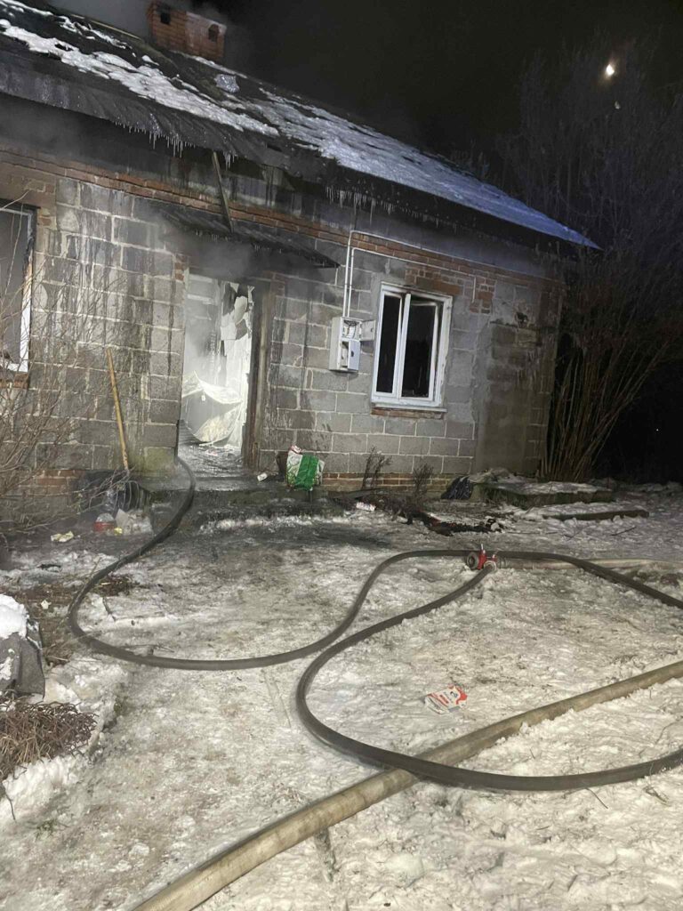 Zniszczony budynek mieszkalny z dymem wydobywającym się z wnętrza, otoczony śniegiem i wężami strażackimi.