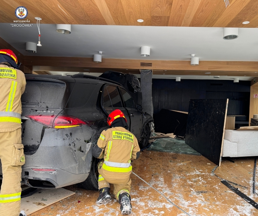 Dwaj strażacy w mundurach przeszukują wnętrze budynku po wjeździe samochodu do środka.