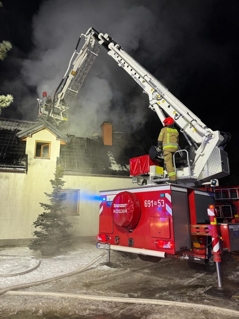 Strażak na podnośniku gaśniczym polewa dach płonącego budynku, w tle widoczny dym i śnieg.