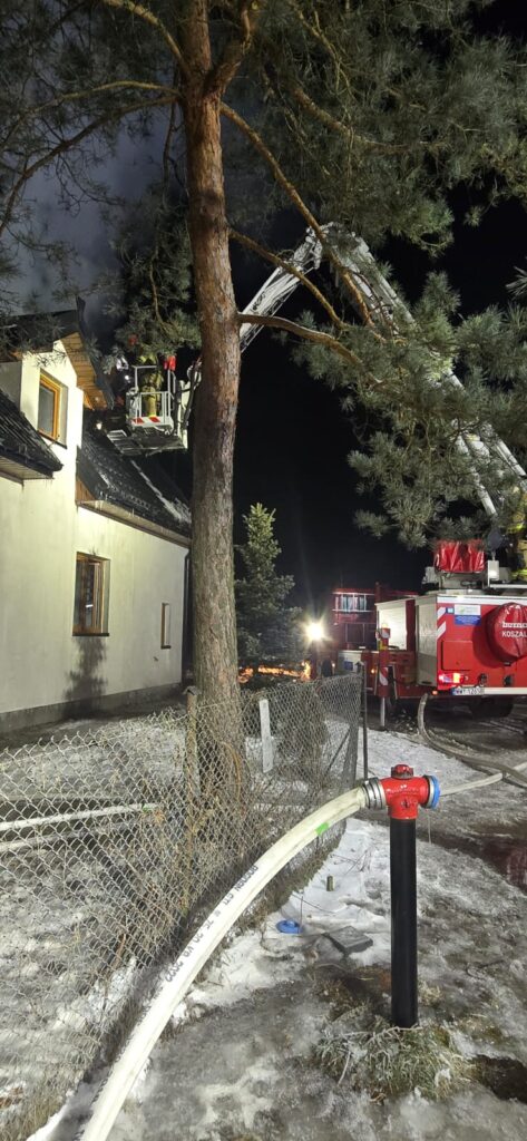 Strażacy na podnośniku gaszą pożar dachu budynku w nocy, otoczeni przez drzewa i sprzęt gaśniczy.