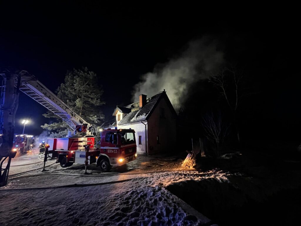 Strażacy gaszą pożar dachu budynku mieszkalnego w nocy, używając wozu strażackiego z podnośnikiem.