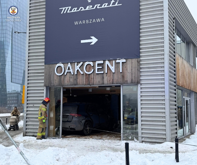 Strażak w mundurze przy uszkodzonej witrynie salonu samochodowego, gdzie wjechał samochód. W tle śnieg i inne budynki.