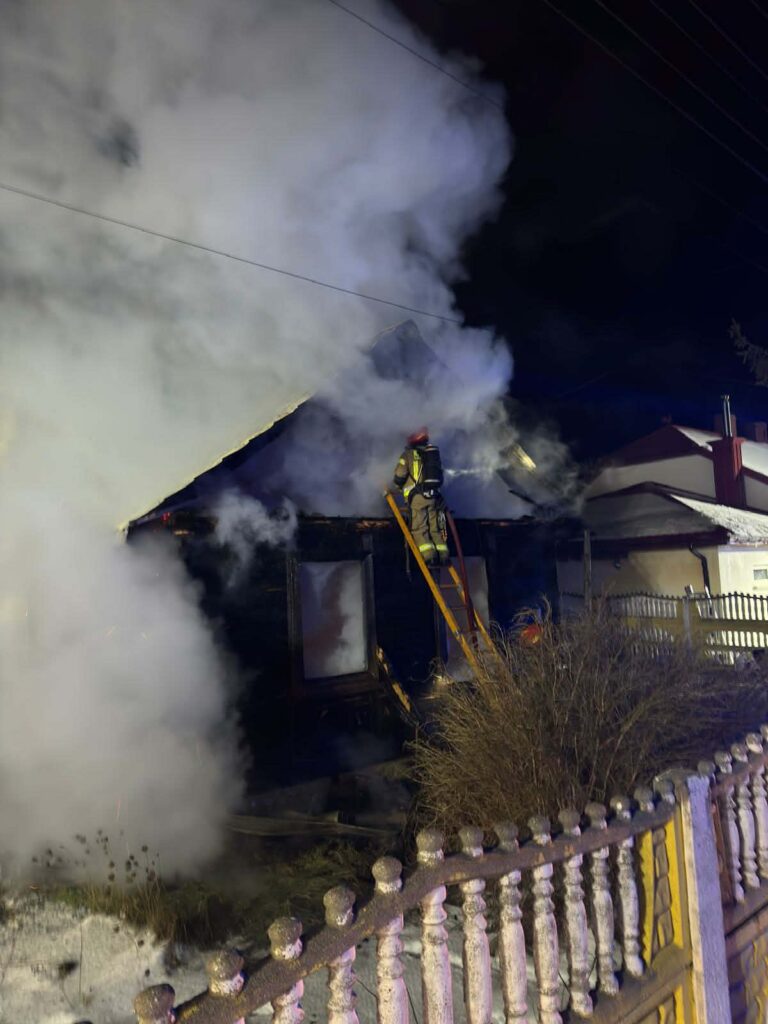 Strażak na drabinie gasi pożar w zadymionym budynku mieszkalnym w nocy. W tle widoczna płot i inne budynki.