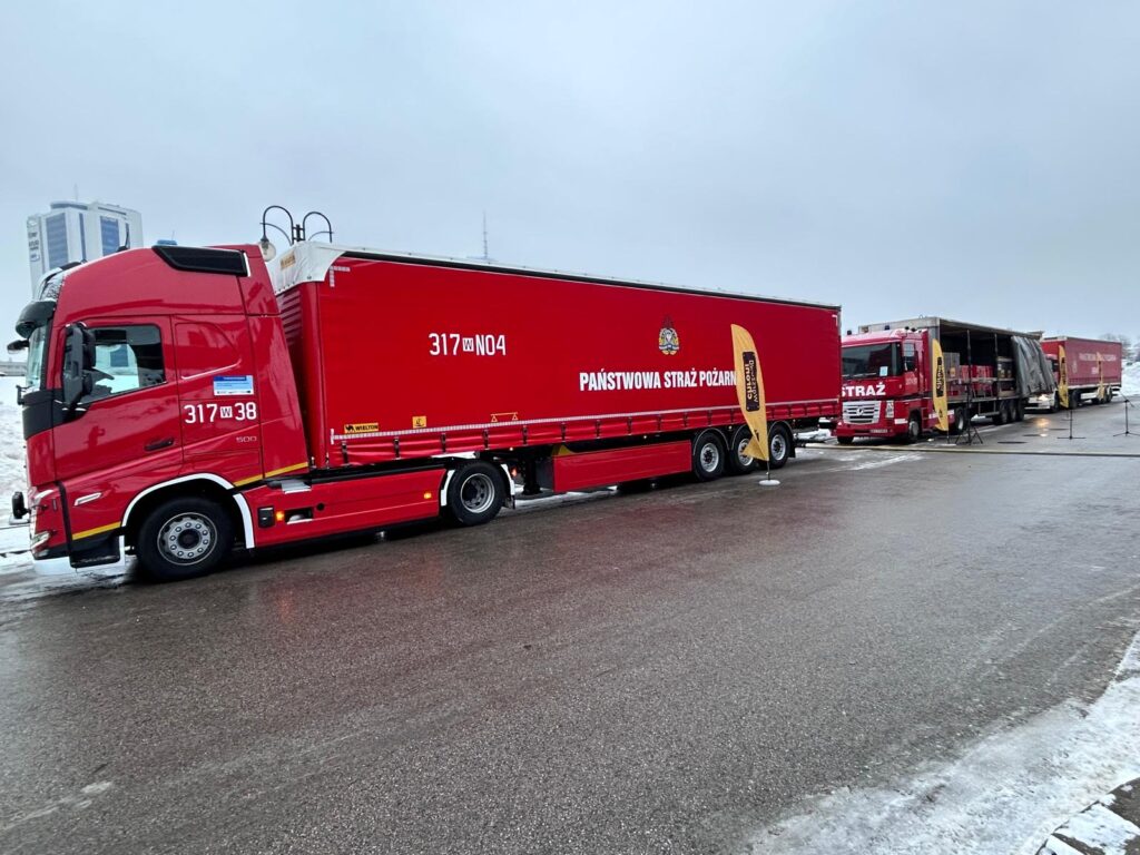 Czerwony samochód ciężarowy z logo Państwowej Straży Pożarnej stojący na parkingu, obok drugi woz z otwartą naczepą.