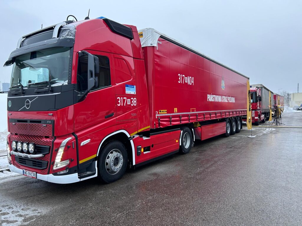 Czerwony samochód ciężarowy z logo Państwowej Straży Pożarnej zaparkowany na placu, obok innych pojazdów.