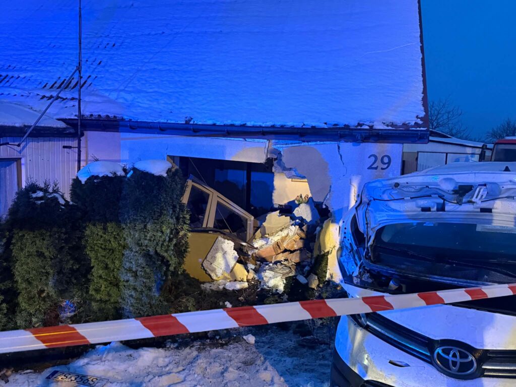 Uszkodzona ściana budynku mieszkalnego po kolizji z samochodem, wokół taśma ostrzegawcza, w tle śnieg i niebo o zmierzchu.