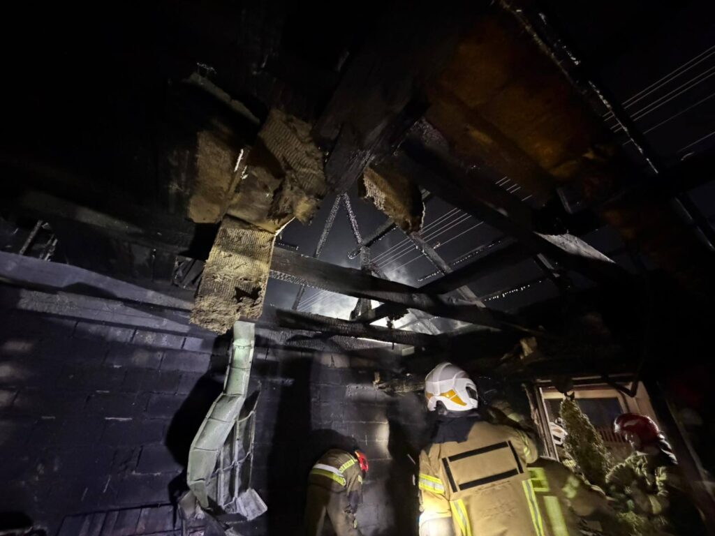 Strażacy pracują w pomieszczeniu z uszkodzoną strukturą dachu, otoczeni przez czarne ściany pokryte sadzą.