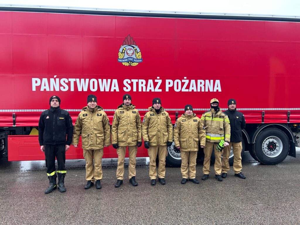 Grupa strażaków w mundurach stoi przed dużą czerwoną ciężarówką z napisem „PAŃSTWOWA STRAŻ POŻARNA”.