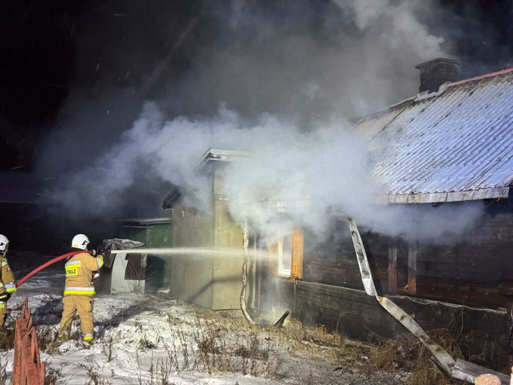 Strażacy gaszą pożar drewnianego budynku, otoczonego gęstym dymem, w zimowej scenerii.