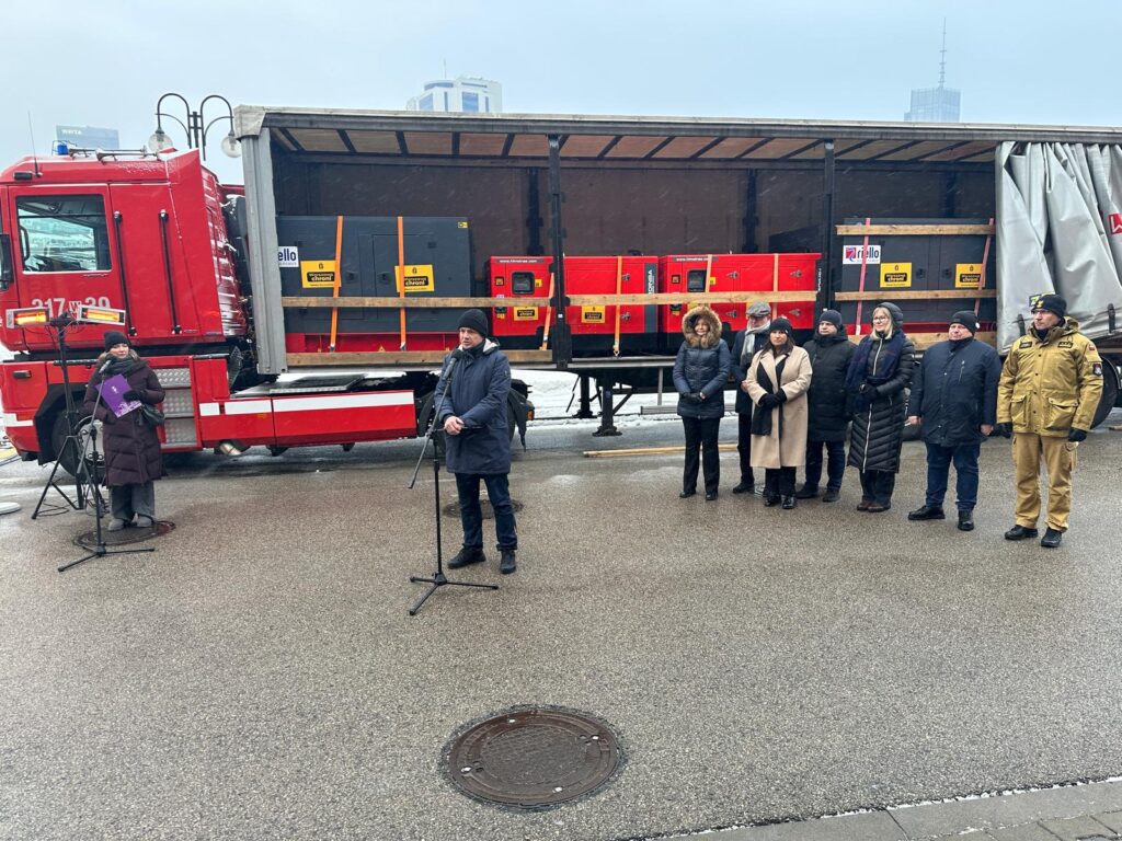 Mężczyzna w garniturze przemawia do mikrofonu, a obok stoi grupa osób w zimowych ubraniach, za nimi ciężarówka z ładunkiem.