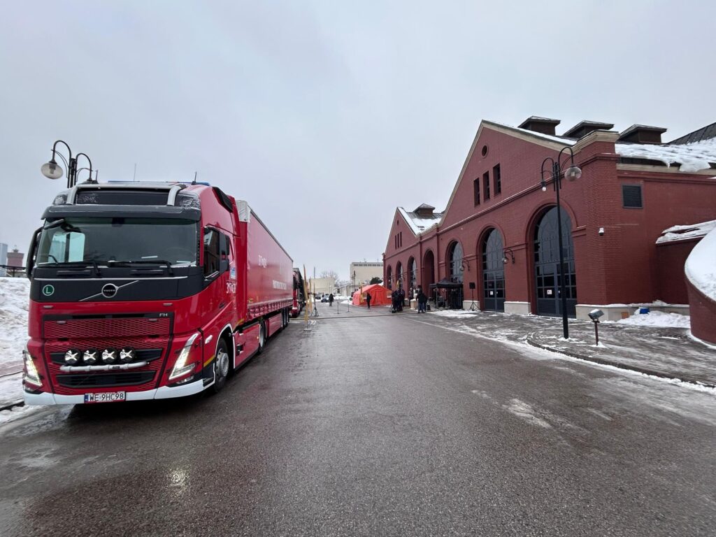 Czerwony ciężarowy samochód zaparkowany na ulicy przed ceglanym budynkiem, obok stoiska z namiotem.