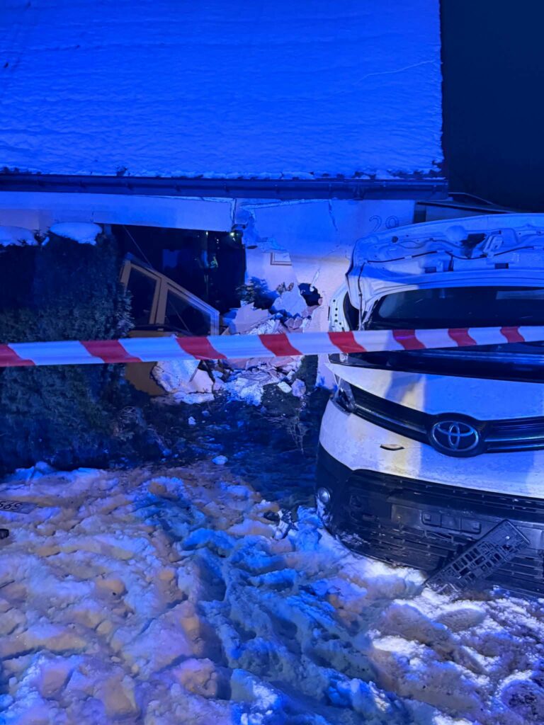 Uszkodzony budynek z widocznym wgnieceniem, w tle zarys samochodu osobowego na zaśnieżonym terenie, otoczony taśmą policyjną.