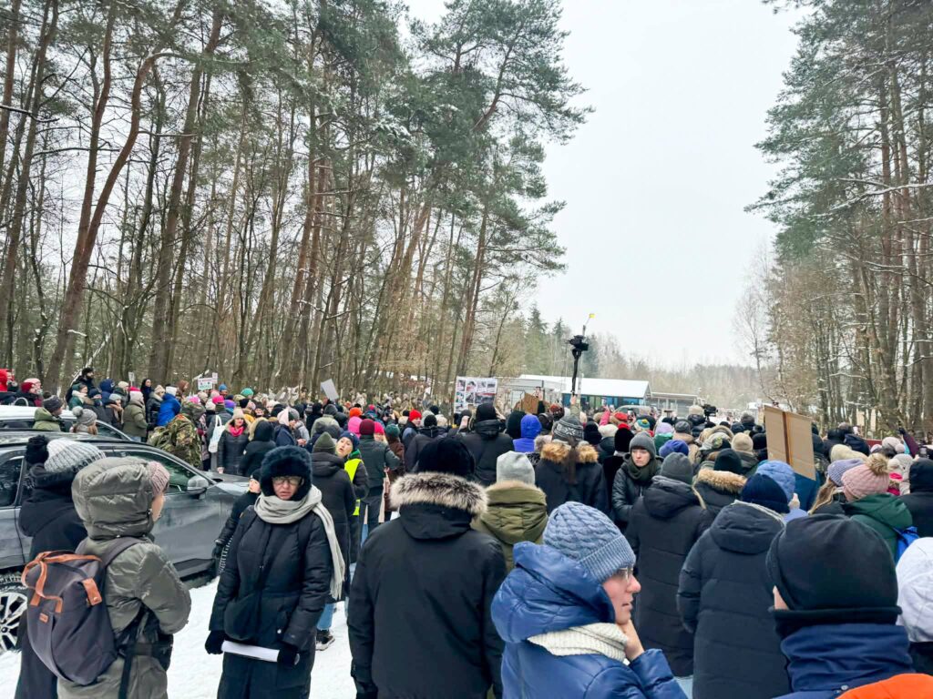 Tłum ludzi w ciepłych ubraniach protestuje w lesie, trzymając transparenty w zimowej scenerii.