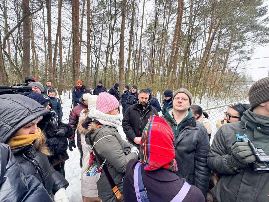Grupa ludzi w zimowych ubraniach stoi w lesie, niektórzy trzymają kamery, w tle widać policyjnych funkcjonariuszy.