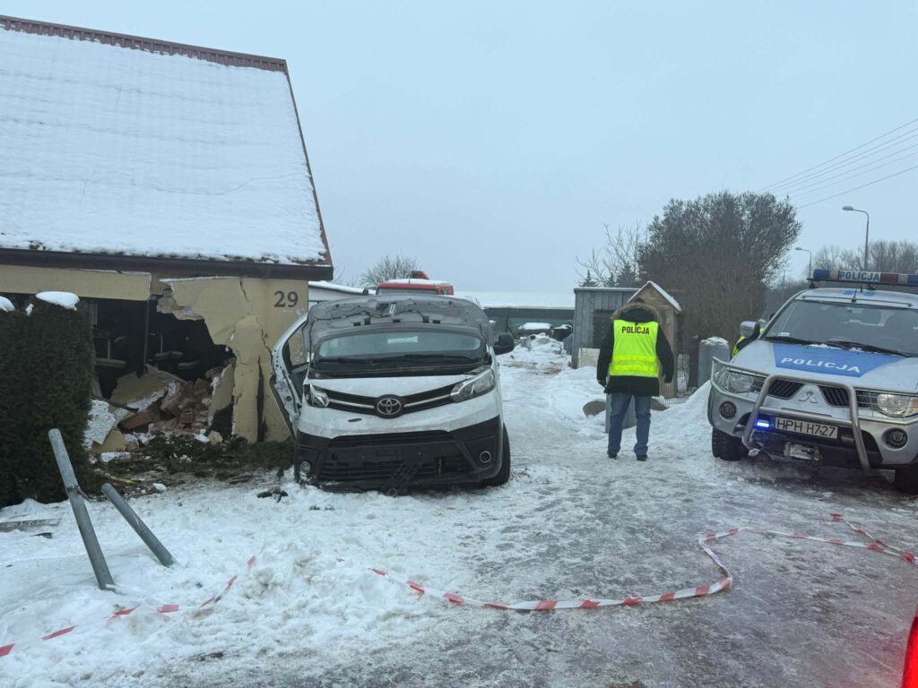 Samochód osobowy uderzył w ścianę domu, gdzie widoczne są zniszczenia, a obok stoi policjant w kamizelce odblaskowej.