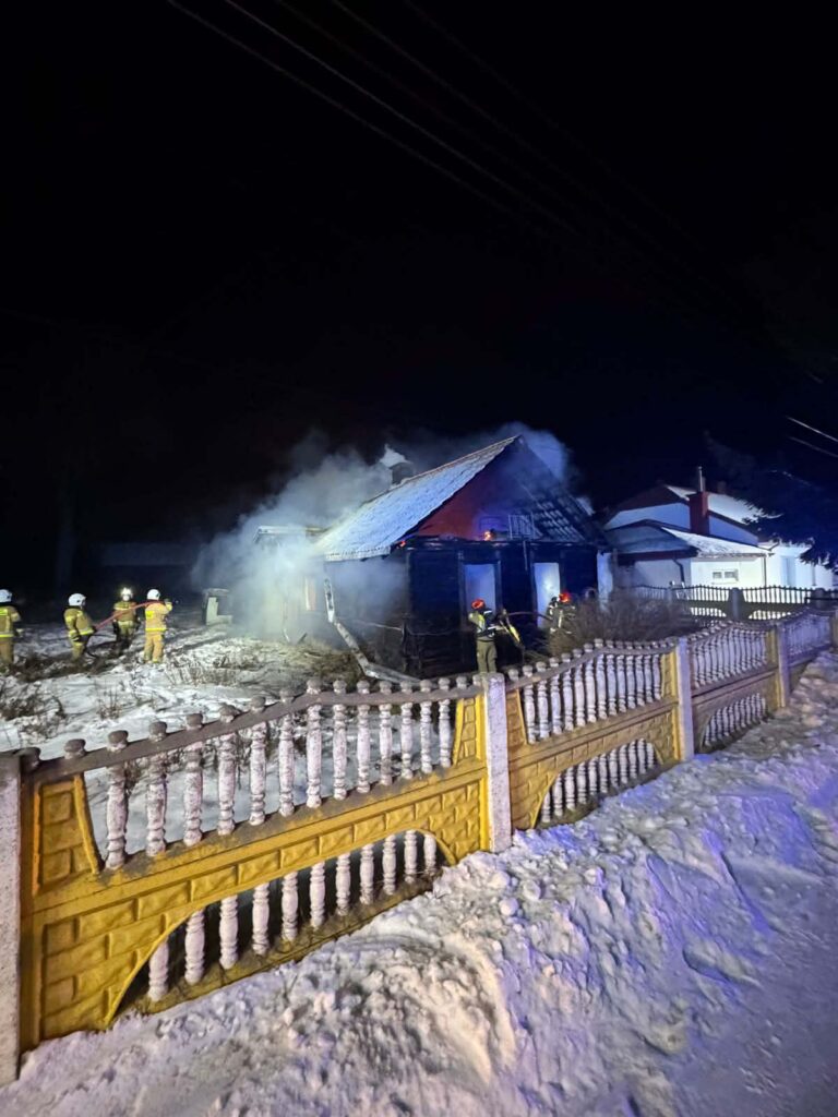 Strażacy gaszą pożar drewnianego domu w zaśnieżonej okolicy nocą, otoczeni dymem.