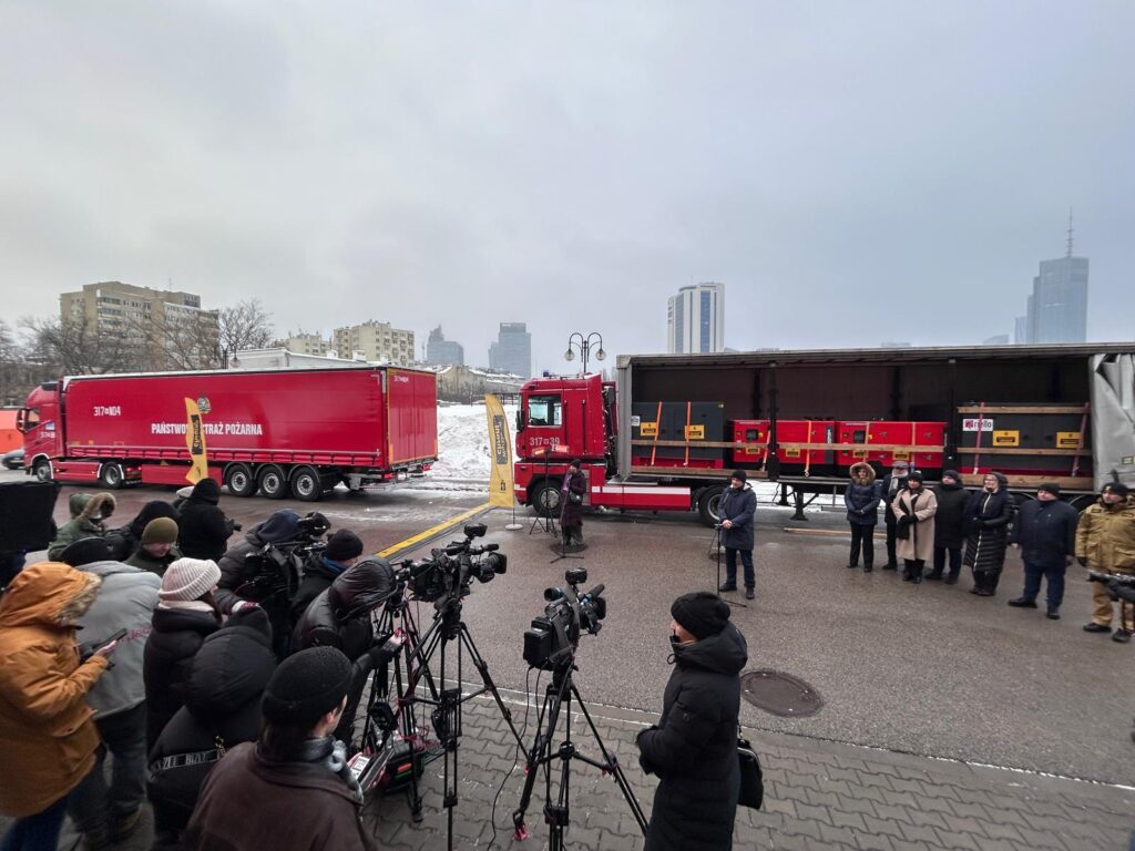 Grupa dziennikarzy z kamerami i mikrofonami stoi przed ciężarówką straży pożarnej, w tle nowoczesne budynki.