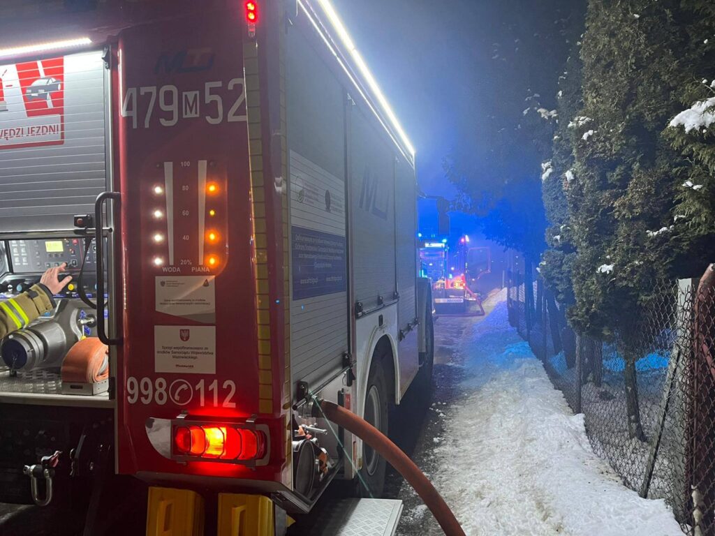 Czerwony wóz strażacki z widocznym panelem sterującym stoi na ośnieżonej drodze, otoczony przez dym i niebieskie światła.