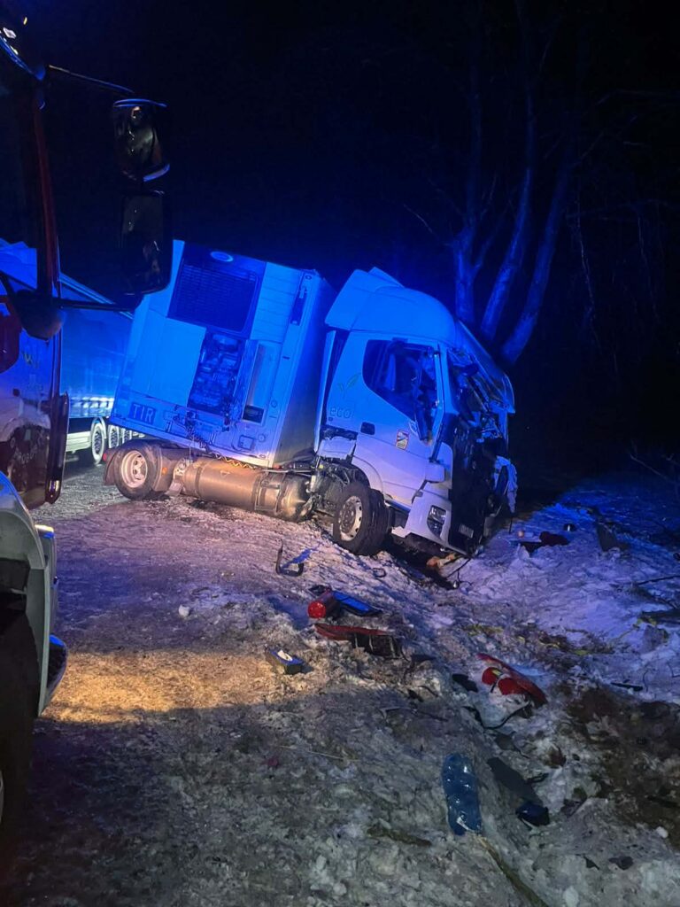 Zniszczona ciężarówka leży na poboczu drogi, w otoczeniu śniegu i gałęzi, w nocy. W tle widoczne niebieskie światła.