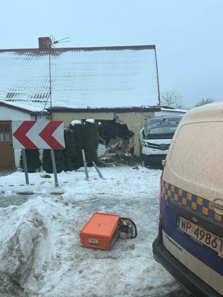 Samochód osobowy wjeżdża w ścianę domu, obok widoczny jest śnieg oraz sprzęt budowlany. W tle oznakowanie drogi.