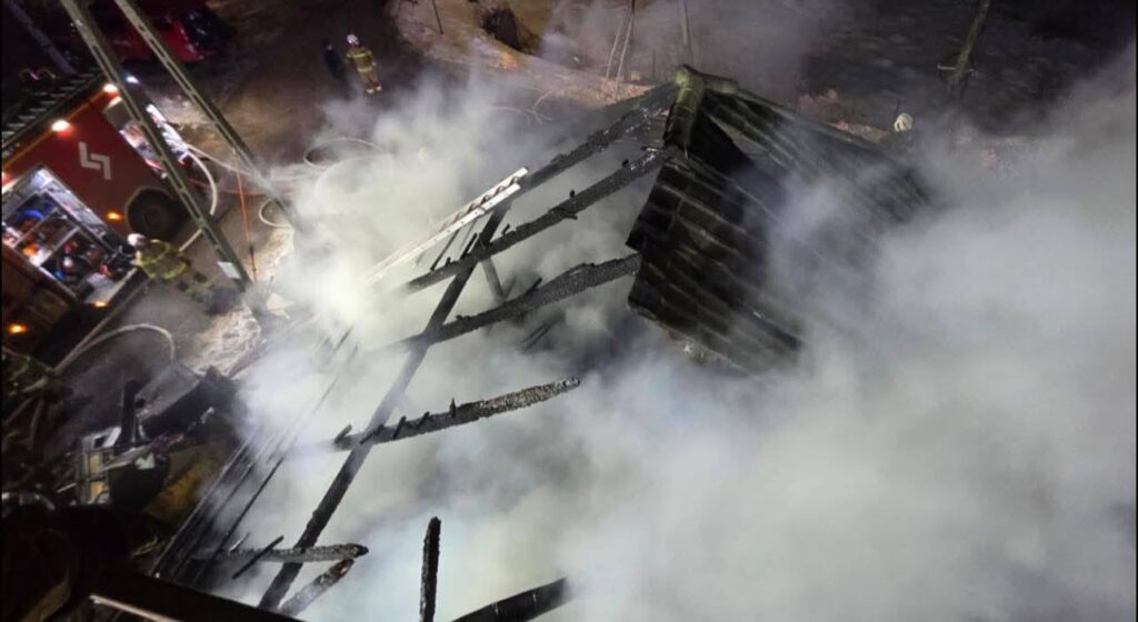 Zniszczony dach budynku pokryty dymem, w tle widać wóz strażacki oraz strażaków gaszących pożar.