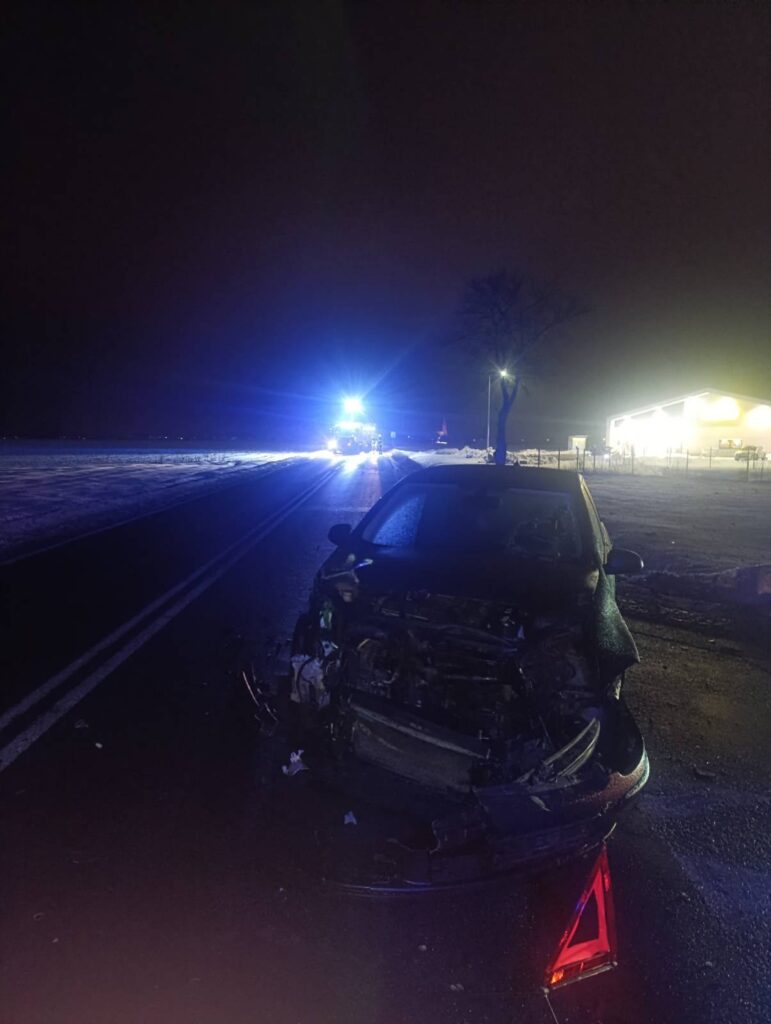 Uszkodzony samochód osobowy stoi na drodze w nocy, w tle widoczne niebieskie światła pojazdu ratunkowego.