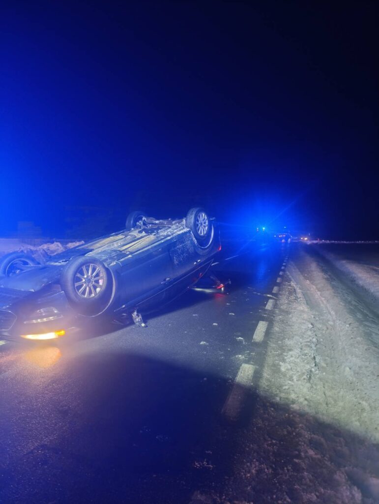 Czarny samochód osobowy leży na dachu na zaśnieżonej drodze, otoczony niebieskim światłem radiowozów.