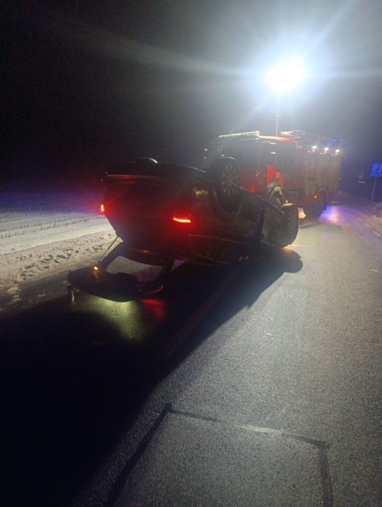 Czarny samochód osobowy leży na dachu na ośnieżonej drodze, w tle widać wóz strażacki z włączonymi światłami.