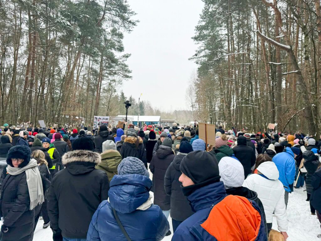 Tłum ludzi w zimowych kurtkach, trzymających transparenty, zgromadzony w lesie podczas demonstracji.