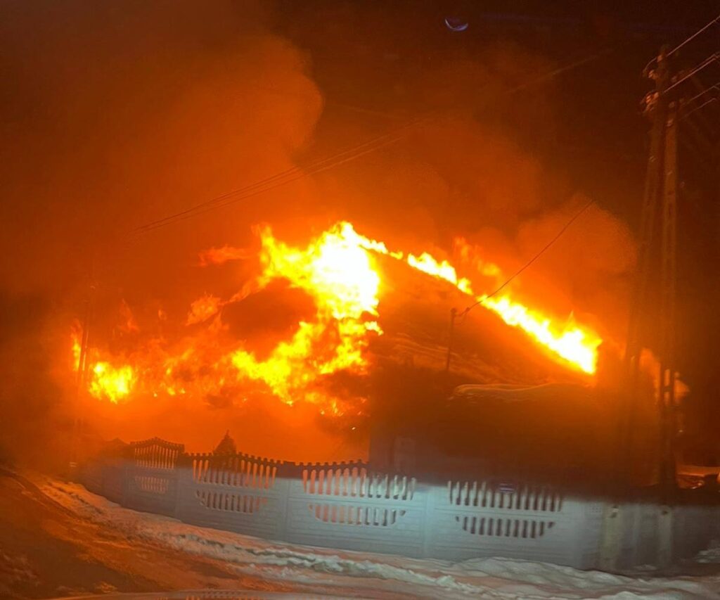 Płonący budynek z intensywnym ogniem i dymem w nocy, w tle widoczny ogrodzenie i słupy energetyczne.