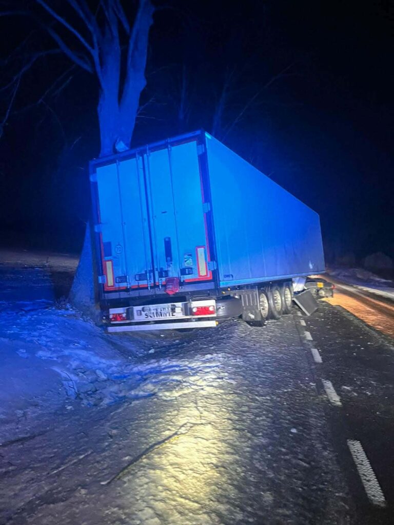 Tir z niebieską naczepą zatrzymany przy drodze, oparty o drzewo w zaśnieżonym terenie nocą.