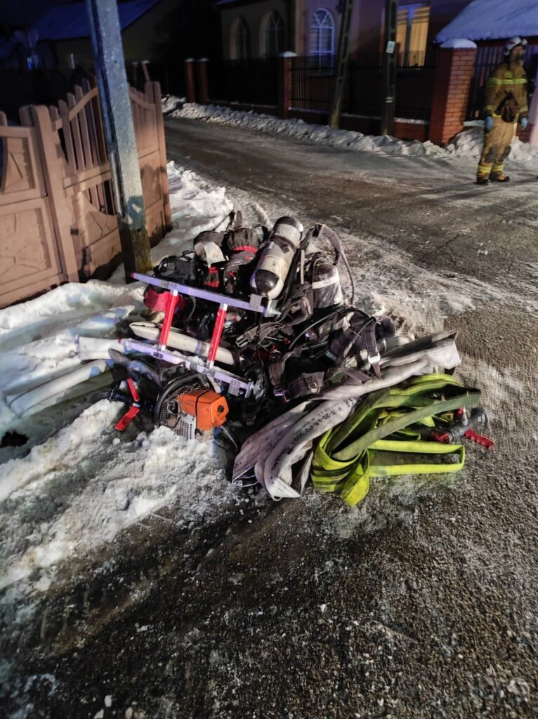 Zestaw sprzętu strażackiego leży na zaśnieżonej drodze obok ogrodzenia, w tle widoczni strażacy.