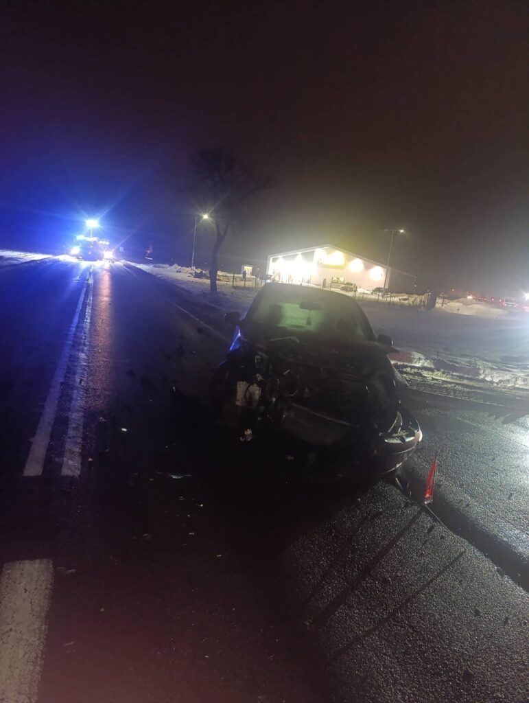 Rozbity samochód osobowy na drodze, w tle włączone światła sygnalizacji i oświetlony budynek.