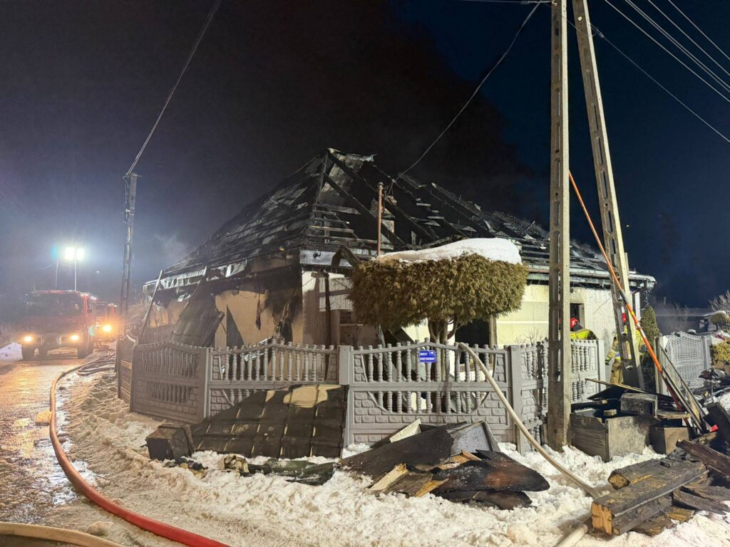 Zniszczony dom z uszkodzoną dachówką, w tle straż pożarna gasząca pożar w nocy, obok leżące resztki drewna.