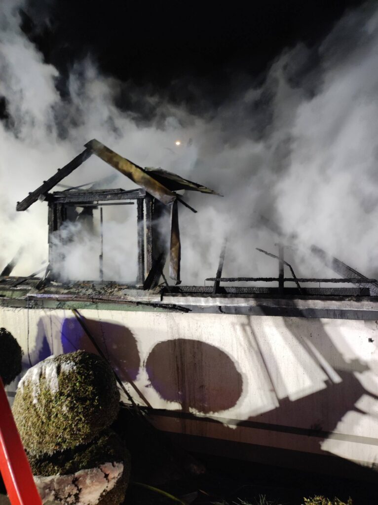 Zgliszcza spalonego budynku pokrytego gęstym dymem, z widocznymi uszkodzeniami dachu i ścian.