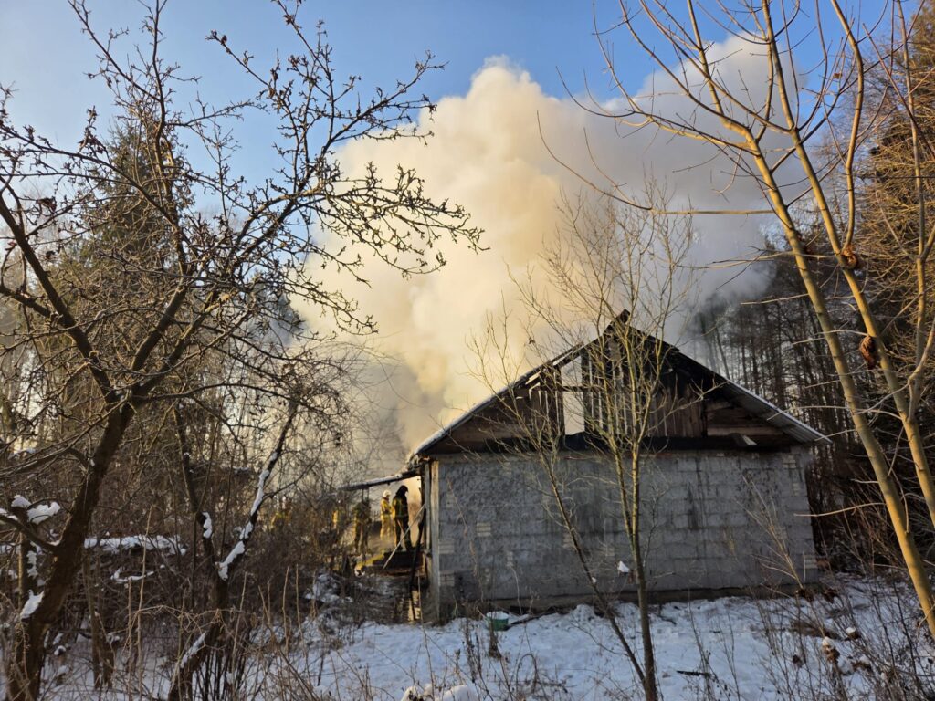 Strażacy gaszą pożar w drewnianym budynku otoczonym śniegiem i drzewami. W tle widać gęsty dym.