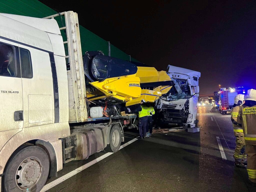 Zderzenie dwóch ciężarówek na drodze, z uszkodzonym pojazdem przewożącym sprzęt budowlany; w tle strażacy i policyjne światła.