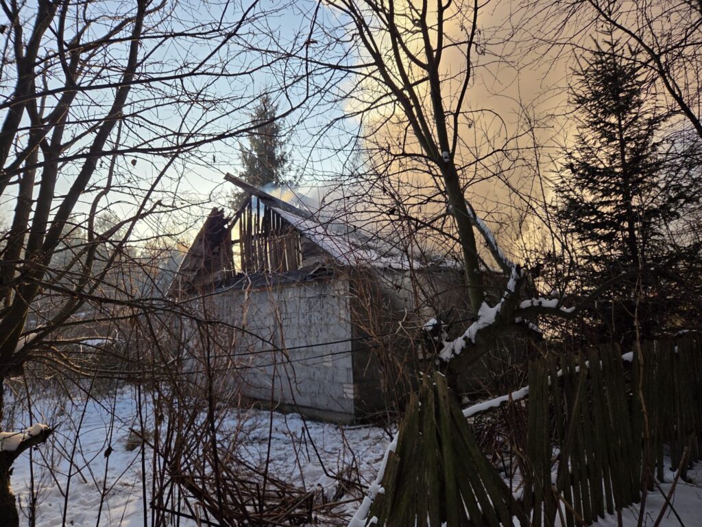 Zniszczony budynek z uszkodzonym dachem, z którego wydobywa się dym, otoczony nagimi drzewami i śniegiem.