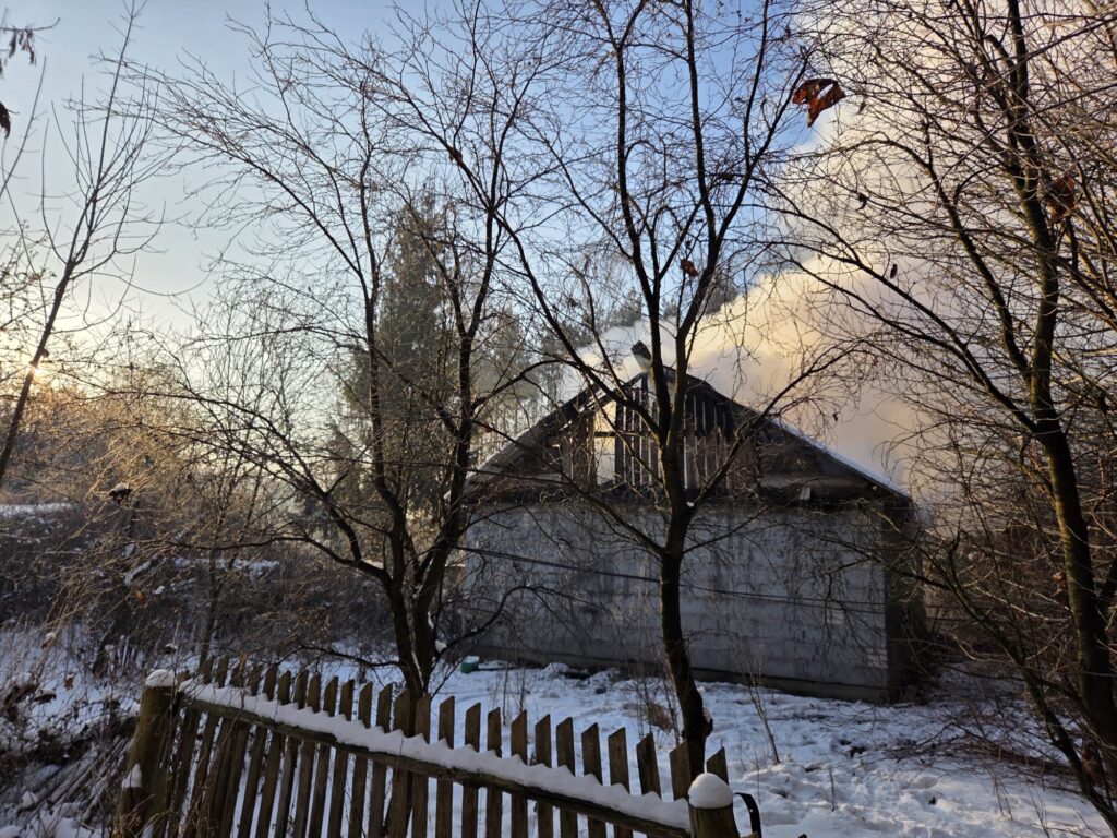 Dym unoszący się z dachu nieukończonego budynku w otoczeniu nagich drzew i pokrytej śniegiem ziemi.