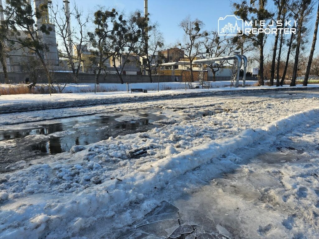 Zamarznięta nawierzchnia pokryta śniegiem i lodem, w tle widoczne drzewa oraz przemysłowe budynki.