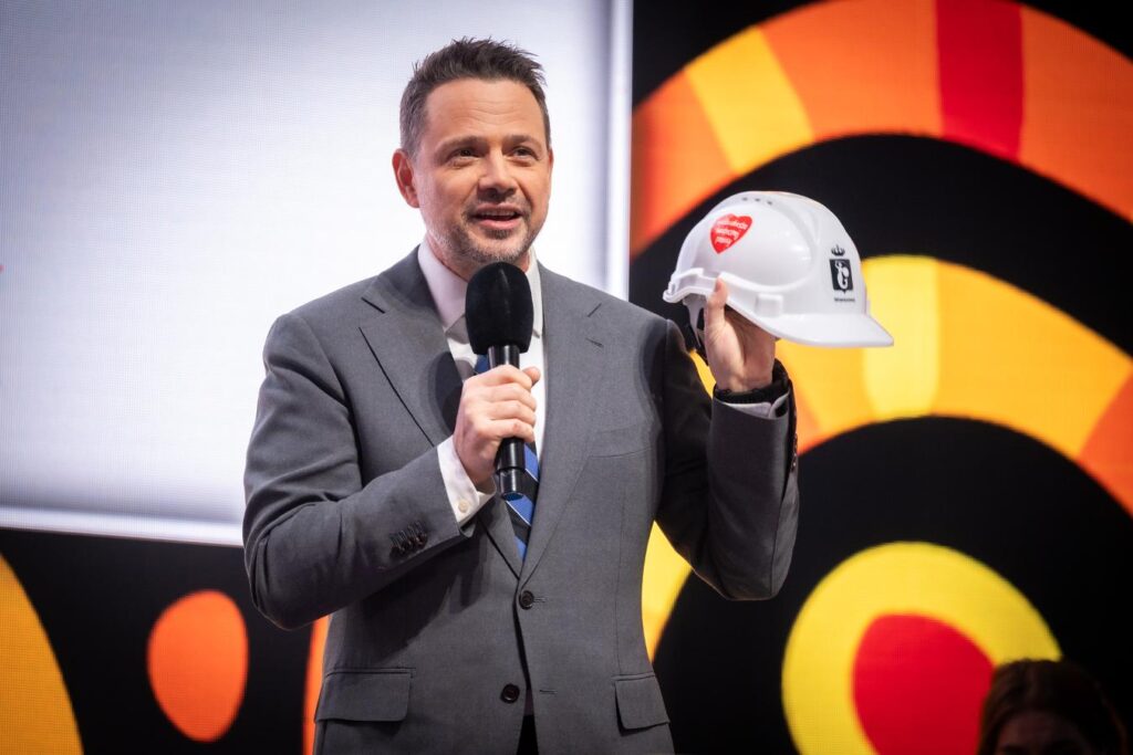 Mężczyzna w garniturze trzyma biały kask budowlany i przemawia do mikrofonu na tle kolorowej sceny.