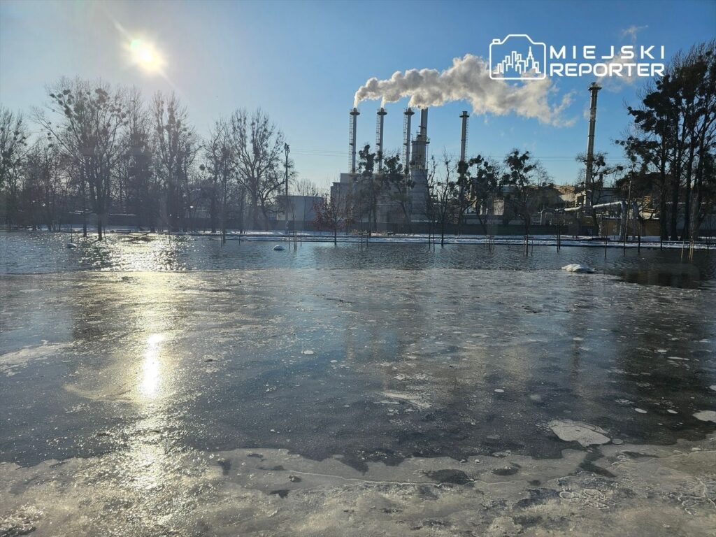 Zamarznięta powierzchnia wody odbija słońce, a w tle widać komin przemysłowy emitujący dym.