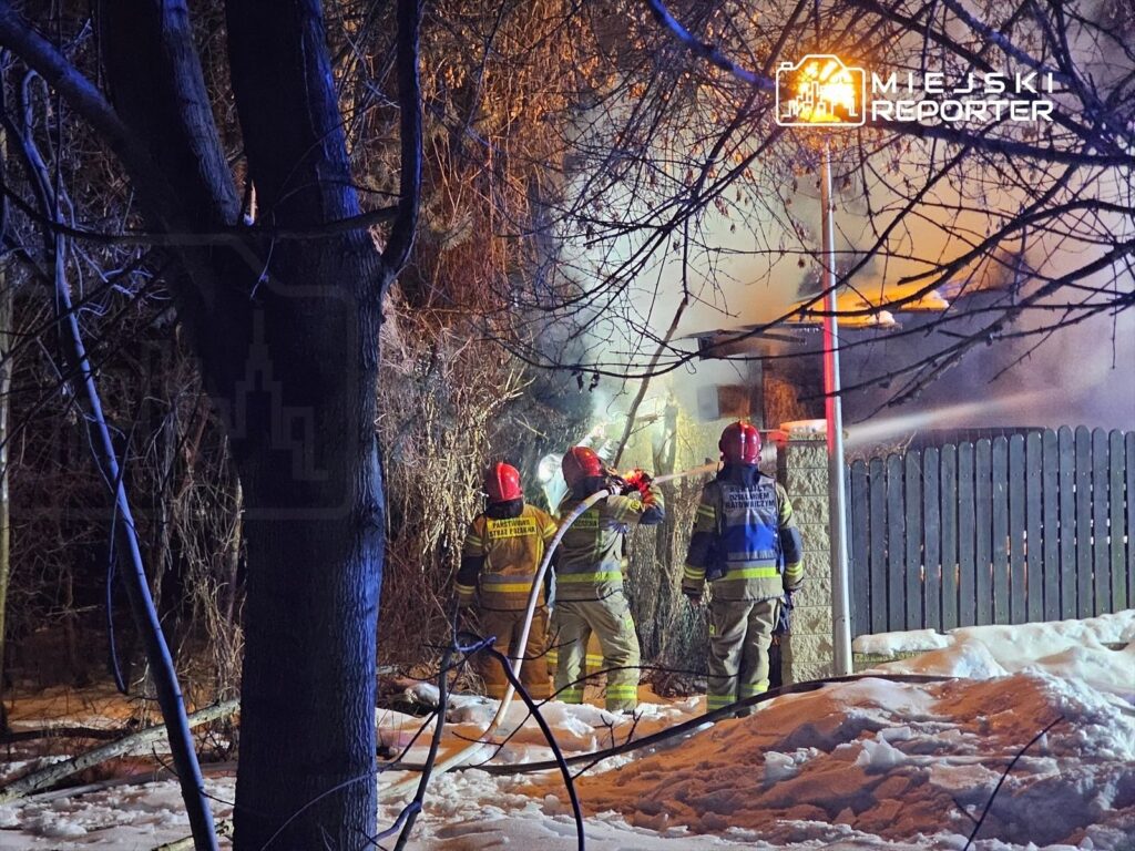 Strażacy gaszą pożar budynku w zimowym otoczeniu, otoczeni przez ośnieżone drzewa i zarośla.