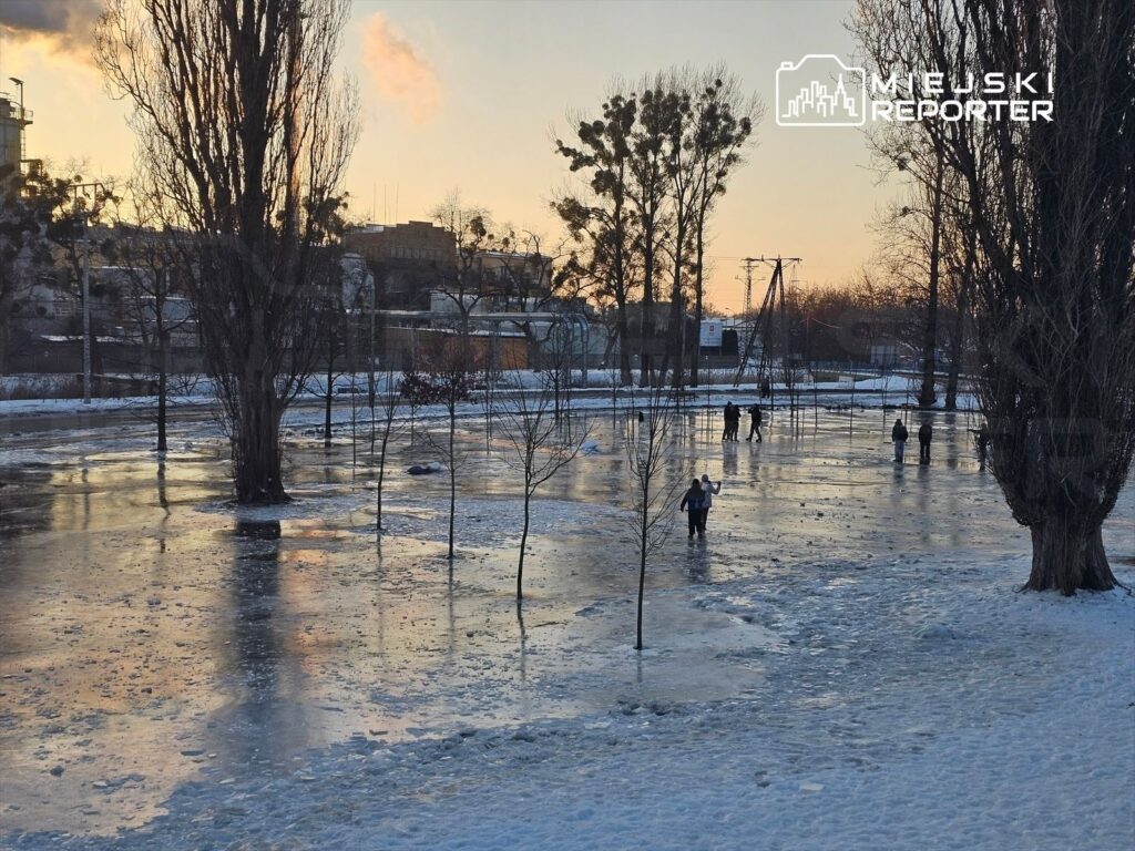 Ludzie spacerują po zamarzniętej powierzchni w parku, otoczeni drzewami i odbijającym się w wodzie zachodem słońca.