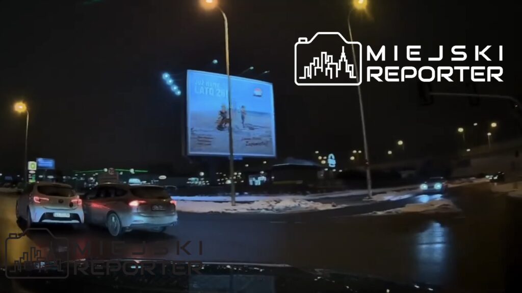 Dwa samochody poruszają się po oświetlonym rondzie w nocy, obok dużego billboardu reklamowego.