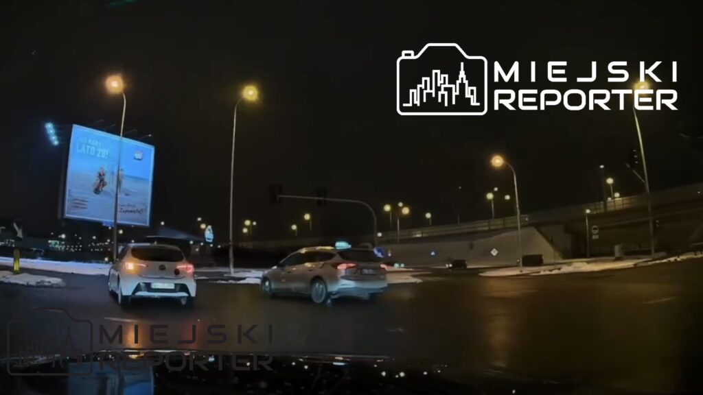 Białe auto osobowe skręca w prawo na rondzie, obok świecący billboard reklamowy w nocy.